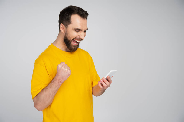excited bearded man with smartphone showing win gesture and shouting isolated on grey - Foto, immagini