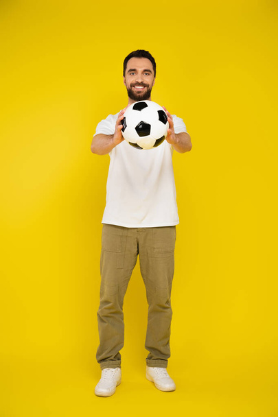 full length of pleased man in pants holding soccer ball in outstretched hands on yellow background - Фото, изображение