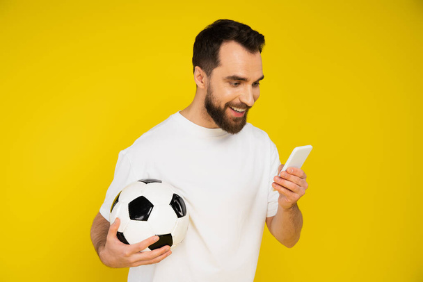 χαμογελαστός οπαδός του ποδοσφαίρου κρατώντας μπάλα ποδοσφαίρου, ενώ βλέποντας το παιχνίδι στο smartphone απομονώνονται σε κίτρινο - Φωτογραφία, εικόνα
