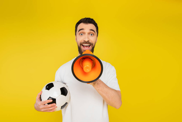 admirador de los deportes sorprendidos con pelota de fútbol gritando en megáfono y mirando a la cámara aislada en amarillo - Foto, imagen