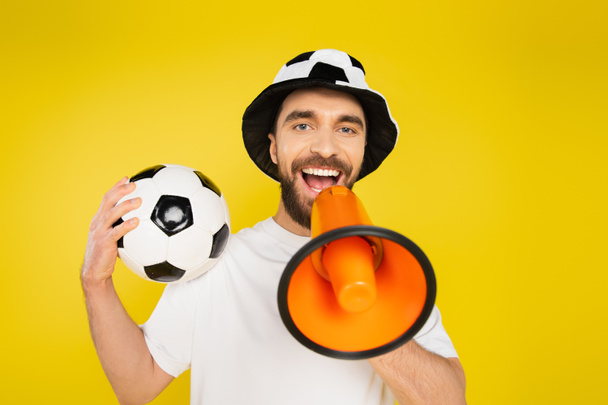 Χαρούμενος άντρας με καπέλο οπαδού του ποδοσφαίρου ουρλιάζοντας σε μεγάφωνο απομονωμένος στο κίτρινο - Φωτογραφία, εικόνα