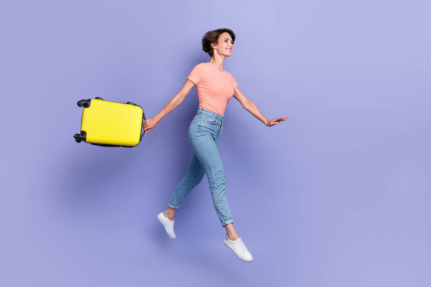 Full length photo of charming sexy girl dressed orange t-shirt jumping walking holding suitcase empty space isolated purple color background. - Fotoğraf, Görsel
