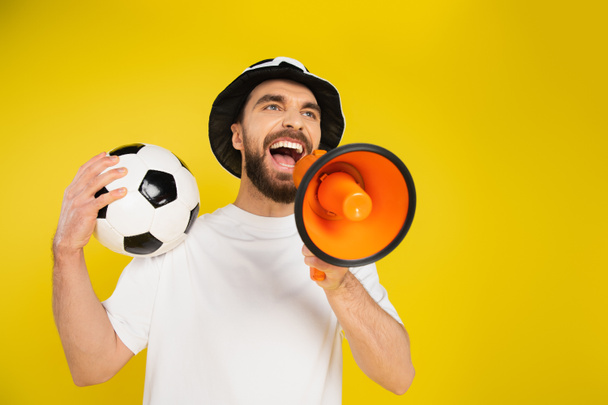 low angle view of sports fan with soccer ball screaming in megaphone isolated on yellow - Foto, afbeelding