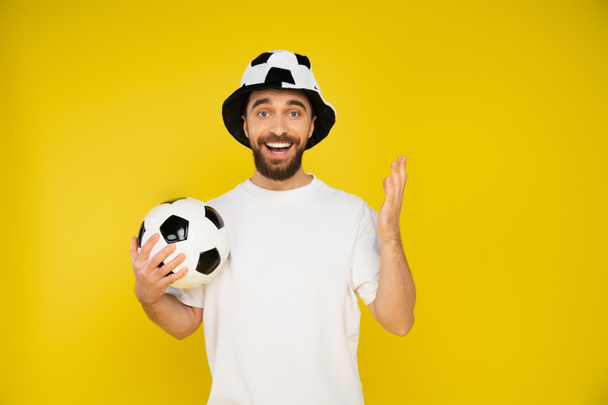 χαρούμενος γενειοφόρος άνδρας στο ποδόσφαιρο καπέλο ανεμιστήρα κρατώντας μπάλα ποδοσφαίρου και κοιτάζοντας κάμερα απομονώνονται σε κίτρινο - Φωτογραφία, εικόνα