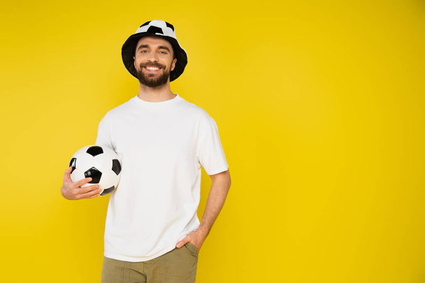 cheerful football fan in hat holding ball and standing with hand in pocket isolated on yellow - Φωτογραφία, εικόνα