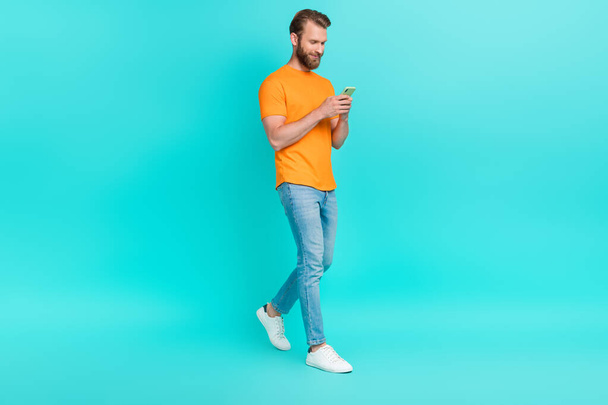 Full size photo of handsome positive man dressed orange t-shirt walking read notification in smartphone isolated on teal color background. - Fotó, kép
