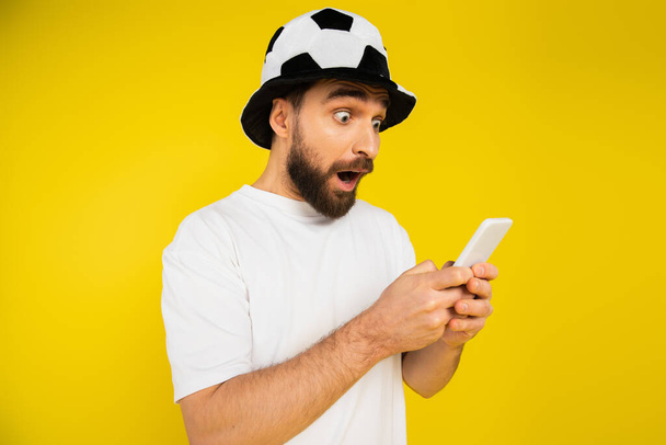 homem chocado no chapéu de fã de futebol assistindo campeonato no celular isolado no amarelo - Foto, Imagem