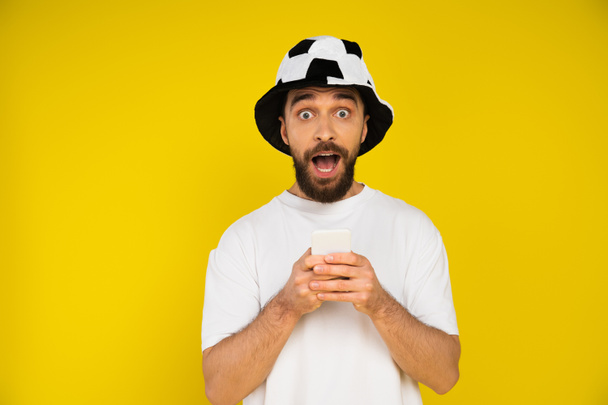 astonished man in football fan hat holding mobile phone and looking at camera isolated on yellow - Photo, Image