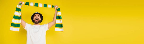 возбужденный спортивный болельщик держа полосатый шарф в поднятых руках и крича изолированы на желтый, баннер - Фото, изображение