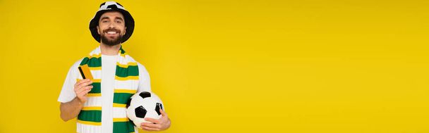 szczęśliwy człowiek w kapeluszu kibica piłki nożnej stojący z karty kredytowej i piłki nożnej izolowane na żółtym, baner - Zdjęcie, obraz