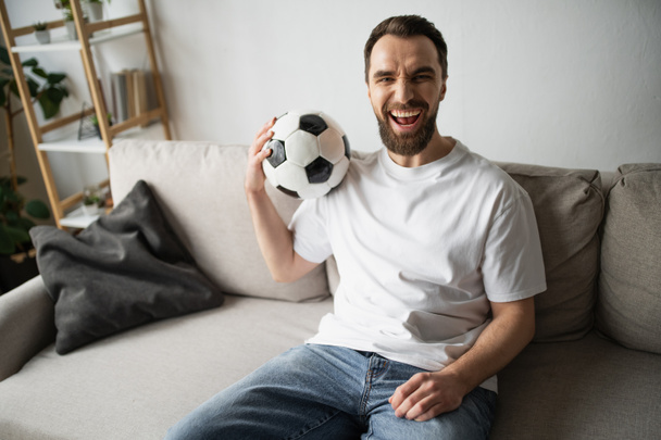 tifoso di calcio eccitato seduto sul divano con pallone da calcio e guardando la fotocamera - Foto, immagini