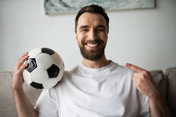 happy bearded man looking at camera and pointing at soccer ball at home - Фото, изображение