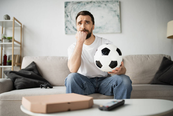 KYIV, UKRAINE - OCTOBER 21, 2022: стурбований чоловік тримає футбол і спостерігає за футболом вдома. - Фото, зображення
