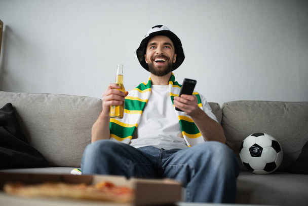 ευτυχισμένος άνθρωπος σε καπέλο ανεμιστήρα και κασκόλ κρατώντας τηλεχειριστήριο και μπουκάλι μπύρα, ενώ βλέποντας ποδόσφαιρο παιχνίδι  - Φωτογραφία, εικόνα