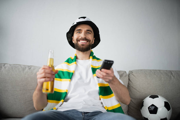 サッカーの試合を見ながらファンの帽子とスカーフで陽気な男がリモコンとビールを持っている  - 写真・画像