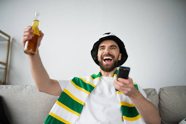 joyful man in fan hat and scarf holding remote controller and bottle of beer while watching football game  - Foto, imagen