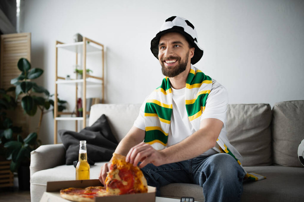 joyful man in hat and scarf taking slice of tasty pizza while watching championship - Photo, Image