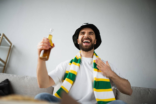 низкий угол зрения изумленного спортивного болельщика в шарфе и шляпе, держащего бутылку пива во время просмотра чемпионата - Фото, изображение