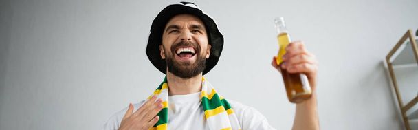 admirador deportivo sorprendido en bufanda y sombrero sosteniendo la botella de cerveza mientras mira el campeonato, bandera - Foto, imagen