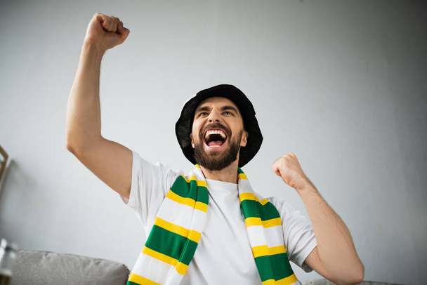 excited man in fan hat and stiped scarf rejoicing and watching championship  - Фото, изображение