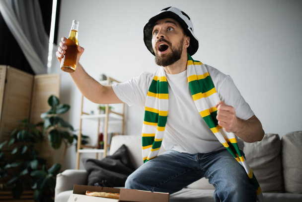 збуджений бородатий чоловік у спортивному капелюсі та шарфі тримає пляшку пива під час перегляду чемпіонату
  - Фото, зображення