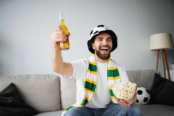 cheerful sports fan in hat and scarf holding bowl with popcorn and bottle of beer while watching championship  - Fotó, kép