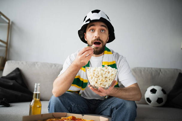 KYIV, UKRAINE - OCTOBER 21, 2022: shocked man in sportive fan hat and scarf eating popcorn and watching championship  - Foto, Bild