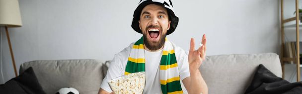 hombre emocionado en el sombrero del ventilador y la bufanda viendo el campeonato mientras sostiene el tazón con palomitas de maíz, bandera - Foto, imagen