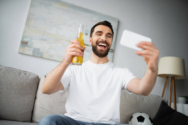 веселый мужчина держит бутылку пива и делает селфи на смартфоне дома - Фото, изображение