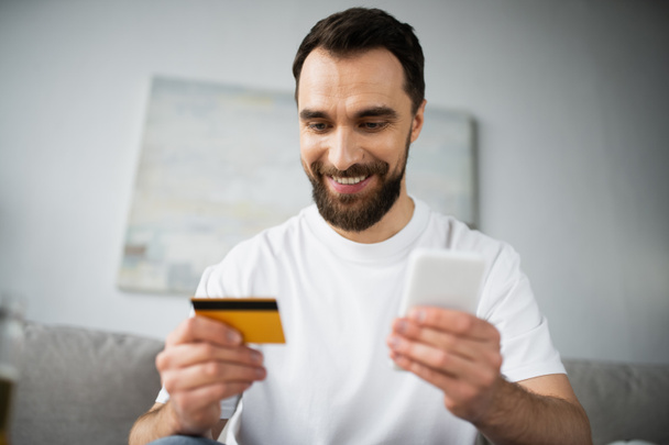 χαρούμενος άνθρωπος που κατέχει πιστωτική κάρτα και τη χρήση smartphone, ενώ κάνει online ψώνια στο σπίτι - Φωτογραφία, εικόνα