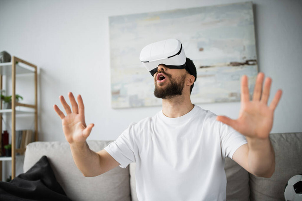 ошеломленный и босый мужчина в гарнитуре VR жестикулирует во время игры дома - Фото, изображение