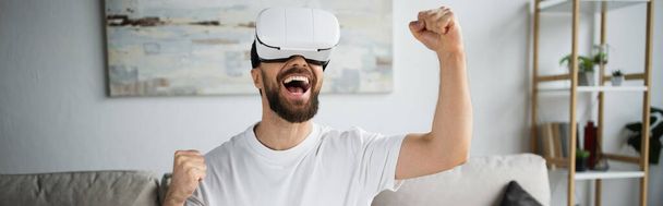 happy and bearded man in vr headset rejoicing while gaming in living room, banner - Foto, imagen