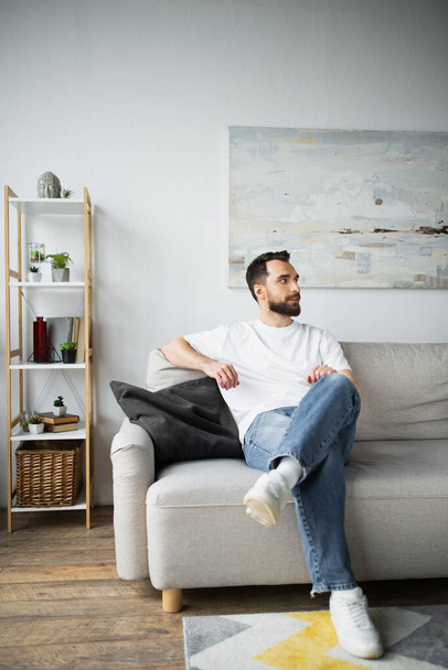 parrakas mies farkut istuu sohvalla modernissa olohuoneessa  - Valokuva, kuva