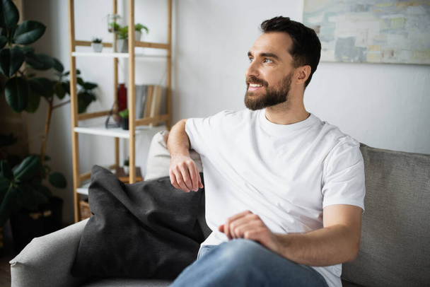 hombre contento y barbudo en jeans sentado en el sofá en la sala de estar moderna  - Foto, Imagen