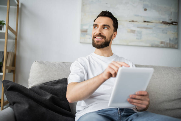 happy man in white t-shirt sitting on couch and using digital tablet in modern living room - Photo, Image