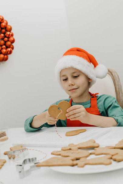 Egy cuki Mikulás sapkás fiú kötelet húzott egy gyömbéres süti lyukába, szív alakban a konyhában, karácsonyi dekorációval díszítve. A karácsonyi ünnepek fogalma - Fotó, kép