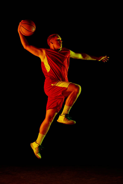 Dynamic portrait of young active athlete, male basketball player in sports uniform practicing with ball, jumping, dribbling isolated over dark background in neon light. Copy space for ad - Foto, afbeelding