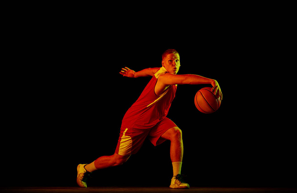 Dribbling. Young man, professional basketball player in action with ball isolated on black background in neon light filter. Concept of sport, energy, skills, team competition. Athlete looks energetic - Foto, afbeelding