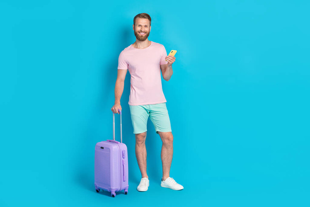 Full body photo of handsome young guy luggage telephone eticket device dressed stylish pink clothes isolated on aquamarine color background. - Fotoğraf, Görsel