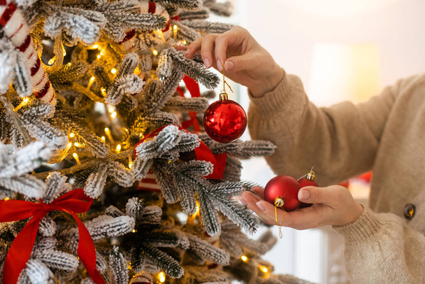 Рождество, зимние праздники и люди - счастливая молодая женщина украшает новогоднюю елку шаром дома. - Фото, изображение