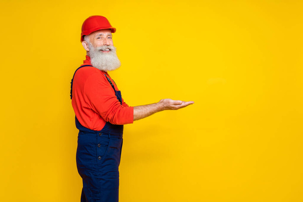 Photo of handsome cute age man workwear overall red hard hat holding arms empty space isolated yellow color background. - Fotografie, Obrázek