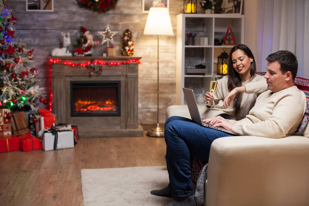 Mooie jonge vrouw en man browsen op laptop voor cadeautjes voor kerstdag. Vrouw met glas champagne. - Foto, afbeelding