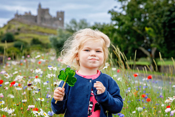 Cute toddler girl with Irish cloverleaf lollipop with Rock of Cashel castle on background. Happy healthy child on flower meadow eating unhealthy sweets. Family and small children vacations in Ireland. - Photo, Image