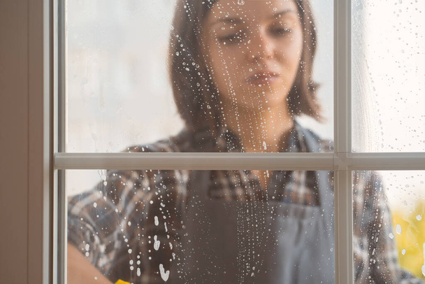 Una joven con guantes de goma amarillos y un uniforme lava las ventanas a fondo con un detergente, secándolas con un trapo, la mujer está limpiando la casa. - Foto, imagen