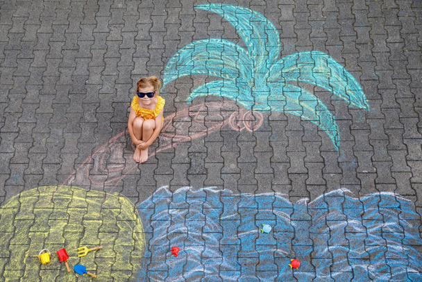 Ευτυχισμένο κοριτσάκι προσχολικής ηλικίας με μαγιό με θάλασσα, άμμο, φοίνικα βαμμένα με πολύχρωμα κιμωλία στην άσφαλτο. Χαριτωμένο παιδί με διασκέδαση με κιμωλία εικόνα. Καλοκαίρι, διακοπές, καλοκαίρι. - Φωτογραφία, εικόνα
