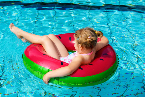 Ευτυχισμένο κοριτσάκι παίζει με πολύχρωμο φουσκωτό δαχτυλίδι στην εξωτερική πισίνα την ηλιόλουστη μέρα του καλοκαιριού. Το παιδί της προσχολικής ηλικίας μαθαίνει κολύμπι. Θερινή υπαίθρια δραστηριότητα. - Φωτογραφία, εικόνα