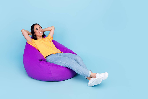 Full size photo of pretty young woman chinese arms behind head sit beanbag dressed stylish yellow clothes isolated on blue color background. - Photo, Image