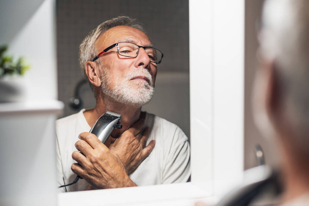 Hombre mayor está recortando su barba con trimmer eléctrico en el baño. - Foto, Imagen