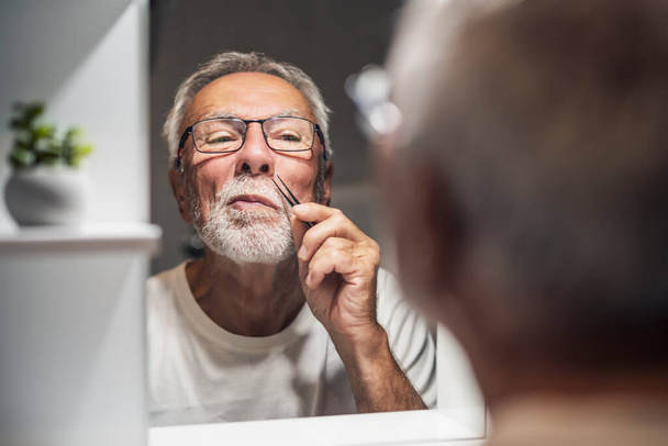 Senior man is picking noise hair with tweezers in bathroom. - Фото, изображение