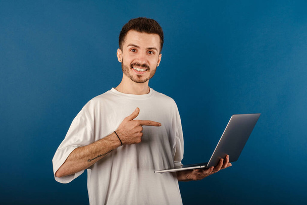 Молодой человек улыбается уверенно одетый в белую футболку позирует изолированно на синем фоне улыбаясь и указывая указательным пальцем на ноутбук компьютера. - Фото, изображение